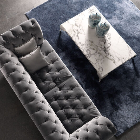 High End Rectangular Marble Coffee Table A top-down view of a modern living room showcases a long, tufted gray sofa paired with a dark blue rug. The High End Rectangular Marble Coffee Table rests elegantly on the rug, adorned with two blue and white porcelain vases. A silver satin pillow is stylishly placed on the sofa.