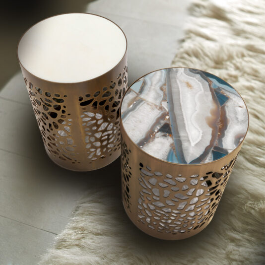 A photograph of two cylindrical side tables showcasing intricate, lace-like laser cut metal sides. The table on the left has a plain white top, while the Italian Designer Laser Cut Bronzed Metal Marble Side Table on the right features a striking multicolored agate-patterned top. Both sit on a cream-colored, fluffy rug.