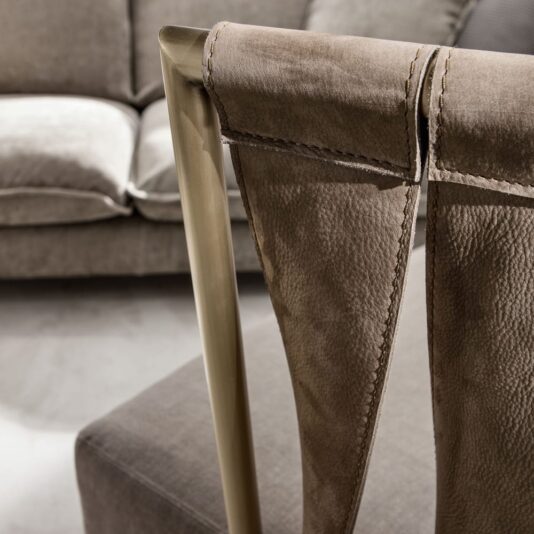 Luxury Italian Designer Leather Twist Armchair Close-up of the back of a Luxury Italian Designer Leather Twist Armchair showcasing its brass frame and light brown leather upholstery. In the background, a brown fabric sofa is visible. The focus remains on the chair’s textures and materials, underscoring its elegant design and superior craftsmanship.