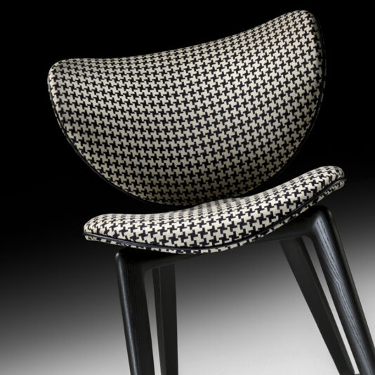 The Designer Retro Occasional Chair features a curved backrest and seat, upholstered in a distinctive black and white houndstooth pattern. Its sleek, black metal legs enhance its contemporary design. The backdrop offers a smooth gradient from dark to light gray, adding to its stylish appeal.