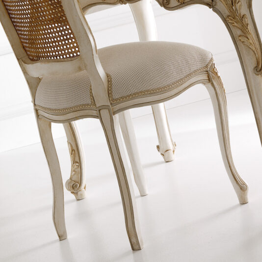 The Italian Designer Rococo Rattan Chair, featuring a beige cushion and intricate carvings, is beautifully showcased in a bright room with a white floor. This vintage-style wooden chair has a refined cane backrest and whitewashed finish, displaying ornate Rococo detailing on the legs and frame.