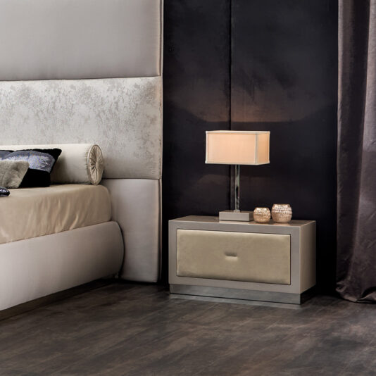 Italian Nubuck Leather Bedside Cabinet Modern bedroom with a luxurious feel. Features a light-colored upholstered bed, an Italian Nubuck Leather Bedside Cabinet with a drawer, and a sleek table lamp. The wall behind is dark with a matte finish, and the floor is a warm wood tone. Two decorative candles sit on the nightstand.