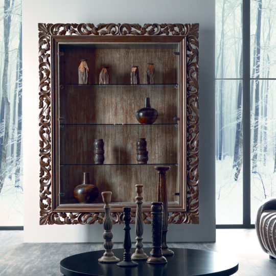An Italian Oak Wall Hanging Display Cabinet with ornate carved edges displays pottery and figures on glass shelves. Wooden candlesticks rest on a round black table in front, with large windows showing a snowy forest beyond.