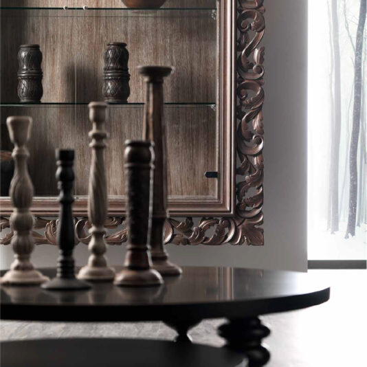 A dimly lit room showcases an intricate Italian Oak Wall Hanging Display Cabinet, adorned with carved detailing and glass shelves. Various wooden candlesticks are arranged on a round table in the foreground, while a large window reveals a snowy forest scene outside.