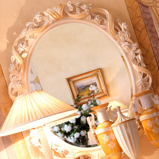 Italian Ornate Oval Reproduction Wall Mirror An Italian Ornate Oval Reproduction Wall Mirror with a floral frame reflects a wall adorned with a painting of a flower vase. Below the mirror, a lamp with a cream-colored shade and two decorative vases add elegance to the setup.