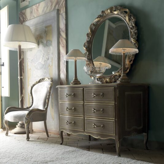 A vintage-style room featuring an ornate wooden dresser with six drawers, adorned with two table lamps and a decorative bowl. Above the dresser hangs the elegant Italian Rose And Ribbon Oval Wall Mirror. A plush, upholstered chair sits to the left, next to a tall floor lamp with a pleated shade.