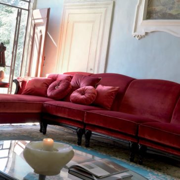 Italian Ruby Red Velvet Sectional Sofa A cozy living room features an Italian Ruby Red Velvet Sectional Sofa, completed with matching plush pillows. Natural light streams through a tall window and an open glass door. A round coffee table in the foreground holds a lit candle and scattered books, enhancing the elegantly decorated space.