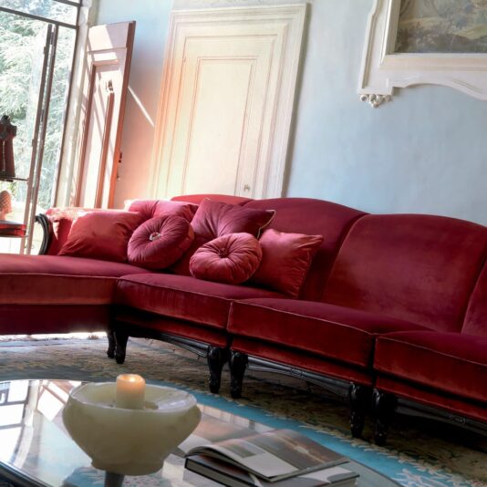 Italian Ruby Red Velvet Sectional Sofa A cozy living room features an Italian Ruby Red Velvet Sectional Sofa, completed with matching plush pillows. Natural light streams through a tall window and an open glass door. A round coffee table in the foreground holds a lit candle and scattered books, enhancing the elegantly decorated space.