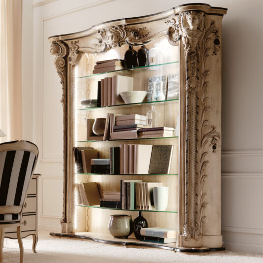 The Large Italian Carved Designer Bookcase, featuring glass shelves and decorative carvings, showcases books, bowls, and vases against a white wall beside a striped chair.