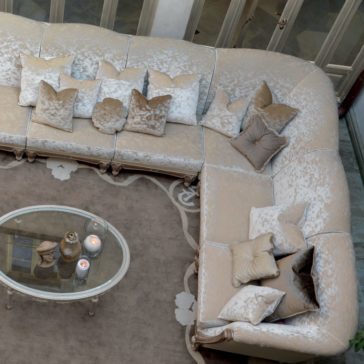 Large Italian Designer Velvet Sectional Sofa A top view of the Large Italian Designer Velvet Sectional Sofa in a plush, silver-grey L-shape, adorned with matching throw pillows in various shades of beige and cream. In front of the sofa is an oval glass coffee table holding lit candles in glass containers and a decorative object.