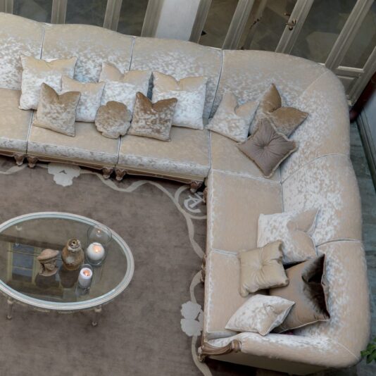 Large Italian Designer Velvet Sectional Sofa A top view of the Large Italian Designer Velvet Sectional Sofa in a plush, silver-grey L-shape, adorned with matching throw pillows in various shades of beige and cream. In front of the sofa is an oval glass coffee table holding lit candles in glass containers and a decorative object.