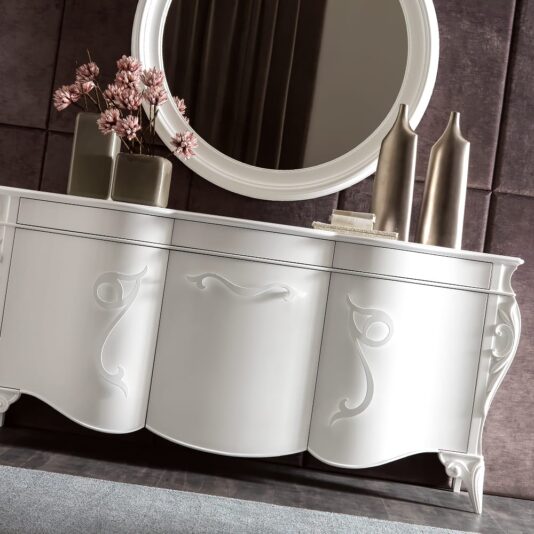 A white, ornate *Italian Elaborate 3 Door Cabinet And Mirror Set* stands against a dark, textured wall. On the cabinet, there is a round mirror, a vase with pink flowers, two tall metallic vases, and a stack of books. The elaborate arrangement adds a touch of elegance and sophistication to the room.