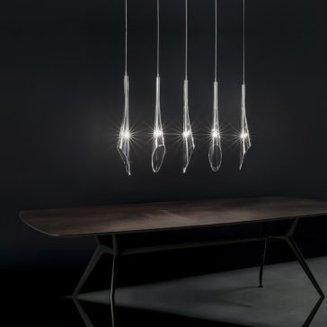 Modern Crystal Glass Rectangular Pendant Light The Modern Crystal Glass Rectangular Pendant Light features five sleek crystal glass elements hanging in a row, casting a radiant glow against a dark backdrop. Below, a wooden table with metal legs enhances the minimalist and elegant setting.