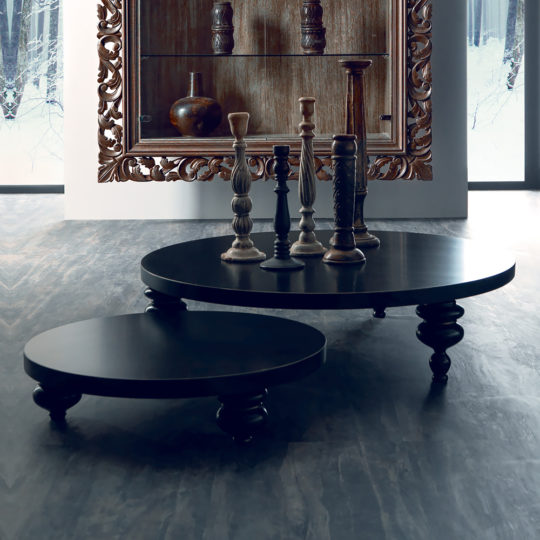 The Low Round Designer Coffee Table Set, featuring two tables of varying heights with ornate legs, stands on a dark floor. Decorative candle holders adorn the tables, and a large ornate mirror is mounted behind them.