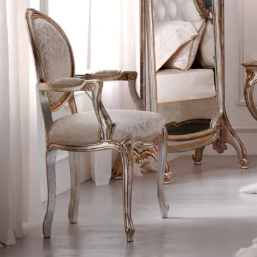 The Luxurious Rococo Upholstered Antiqued Armchair features a patterned fabric seat and backrest, along with elegant wooden carvings adorned with gold accents. This exquisite armchair is placed in a softly lit room next to a bed and matching mirror, enhancing the luxurious and classic decor.
