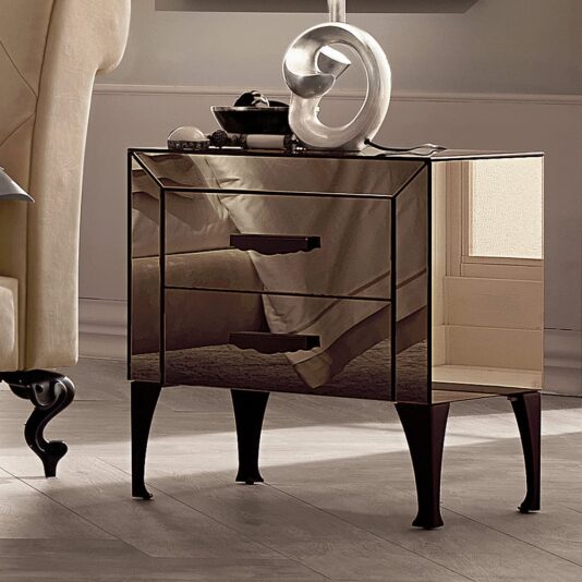 The Luxury Bronzed Mirrored Bedside Table features a modern design with two drawers and sleek black handles. It stands on four black, curved legs and is positioned next to a beige upholstered chair. The table's surface showcases a decorative sculpture and some small items.