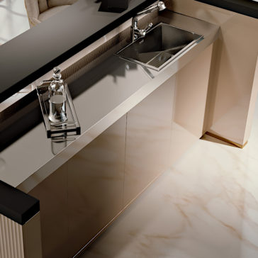 A modern kitchen counter with a shiny, reflective stainless steel surface exudes the elegance of the Luxury High End Italian Cocktail Bar. Nestled on the counter is a polished metal tray holding a glass water bottle and glass. The counter also features a built-in sink and faucet, set above beige cabinet doors.