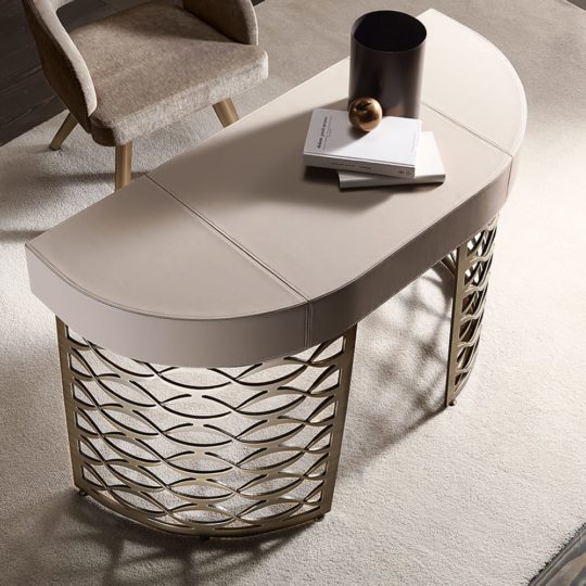 A Luxury Italian Designer Bronze Frame Desk with a smooth beige top and gold metal base is paired with a beige upholstered chair on light carpet. Books, a decorative sphere, and a vase enhance the sophisticated workspace.