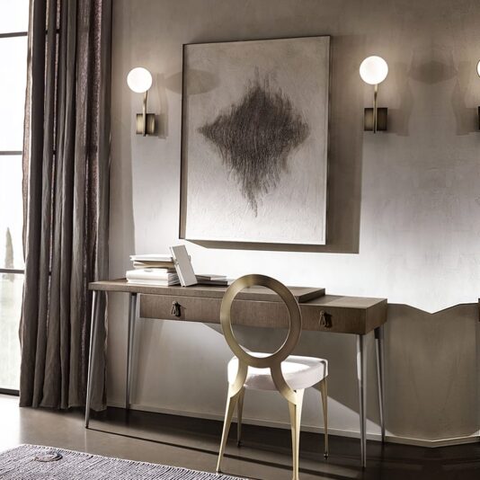 A modern home office features the sleek Luxury Italian Designer Contemporary Desk, adorned with a white laptop and several books. A round-back chair is positioned in front of the contemporary desk. Abstract art and two round wall lamps enhance the decor, while floor-length curtains frame the large window on the left.