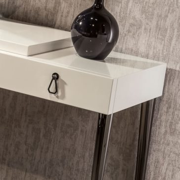 Italian Designer Ivory Lacquered Dressing Table With Mirror A close-up of the Italian Designer Ivory Lacquered Dressing Table with Mirror, showcasing its modern design and single drawer adorned with a black handle. Atop the table sits a decorative glossy black vase. The sleek piece stands on shiny, metallic legs against a textured grey wall backdrop.