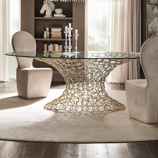 Luxury Italian Rings Glass Dining Table A modern dining room features the Luxury Italian Rings Glass Dining Table, showcasing a sophisticated glass top and an intricate gold wire base inspired by Italian design. Two beige, high-back chairs are positioned on opposite sides of the table. The room is adorned with tall candlesticks, geometric shelves, and neutral tones, creating an elegant ambiance.