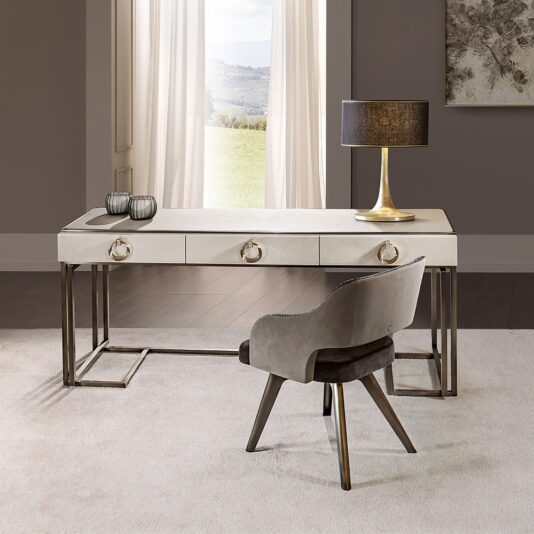 A Luxury Italian Mother Of Pearl Writing Desk with three drawers stands in a bright room with large windows, light curtains, gray walls, artwork, and a gray upholstered chair on a light carpet.