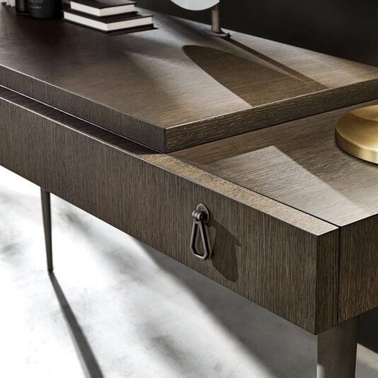 A close-up view of a modern wooden desk with a sleek, minimalist design. Reminiscent of the Luxury Italian Designer Extendable Media Unit, the desk features a metallic handle on one drawer and thin legs, while a lamp and books adorn its smooth, dark-finished surface, creating a contemporary ambiance.