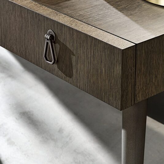 A close-up view of the corner of a wooden table featuring one drawer with a unique teardrop-shaped metal handle. Reminiscent of the Luxury Italian Designer Extendable Media Unit, the table boasts a dark wood finish and smooth surface, with slim legs that are slightly darker in color.