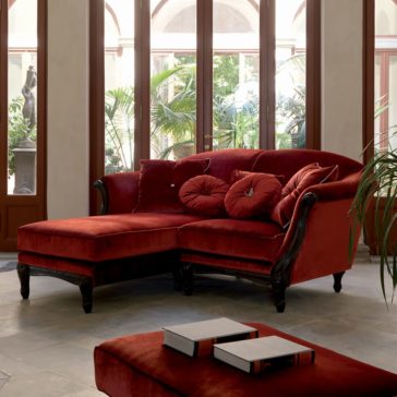 The Luxury Red Velvet Italian Designer Sectional Sofa with round pillows sits in a bright room with large windows and green plants, while a matching ottoman topped with books is placed in the foreground.