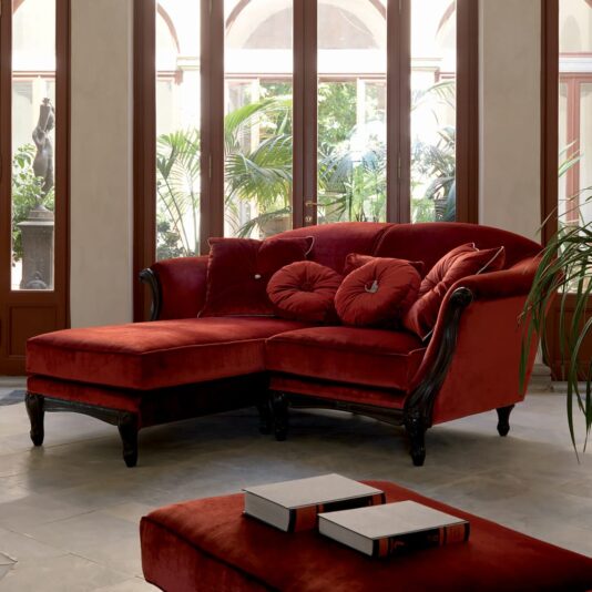 Luxury Red Velvet Italian Designer Sectional Sofa The Luxury Red Velvet Italian Designer Sectional Sofa with round pillows sits in a bright room with large windows and green plants, while a matching ottoman topped with books is placed in the foreground.