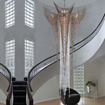 Luxury Stairwell Designer Crystal Pendant Chandelier The Luxury Stairwell Designer Crystal Pendant Chandelier features cascading crystal strands and hangs in a bright entrance hall with curved staircases, glass block windows, and a black chair by a small table topped with flowers.