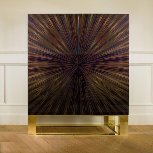 The Modern High End Italian Ebony Veneered Cabinet boasts a striking starburst pattern in various shades of brown and black. Positioned against a beige wall with white molding, this luxurious wooden cabinet stands on a rectangular gold metal base, complemented by the light wood flooring.