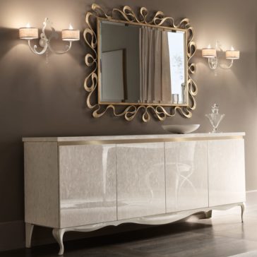 Modern Italian Designer Champagne Leaf Mirror A stylish room featuring a sleek, reflective sideboard with a white finish and curvy legs. Above it, the Modern Italian Designer Champagne Leaf Mirror with its gold decorative frame exudes elegance. This ornate mirror is flanked by two elegant wall sconces, and a white bowl and glass decanter sit on the sideboard.