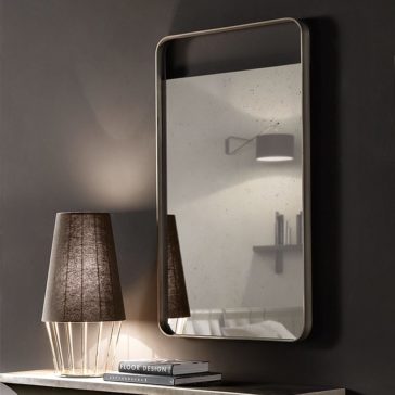 Modern Italian Designer Curved Edge Mirror A minimalist room features a sleek Modern Italian Designer Curved Edge Mirror, mounted on a dark wall. Below the mirror, a modern lamp with a brown shade sits on a table, alongside a neatly stacked book titled
