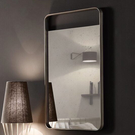 Modern Italian Designer Curved Edge Mirror A Modern Italian Designer Curved Edge Mirror is mounted on a dark wall. Below the mirror, a dark-colored lamp with a textured shade emits light towards the wall. The mirror reflects a partial view of the lamp and a shelf in the room.