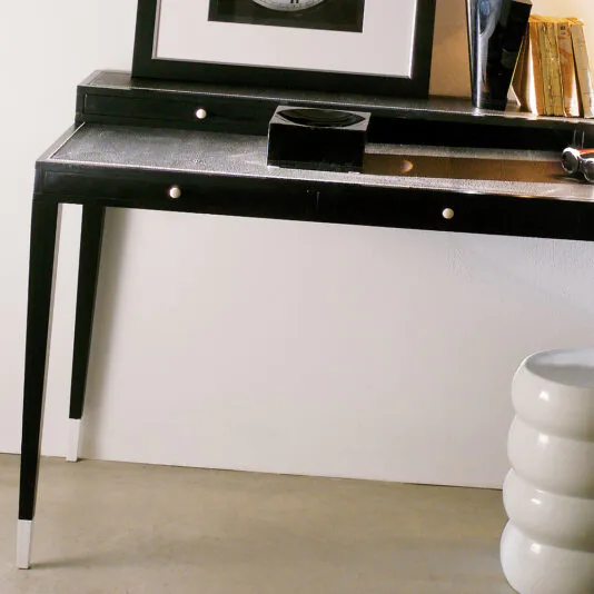 A minimalist room showcasing a Modern Italian Leather Writing Desk with two drawers, accompanied by a small black box and a stack of books on top. A framed picture is mounted on the wall above the desk. On the right, a white, round, three-tiered stool is partially visible.