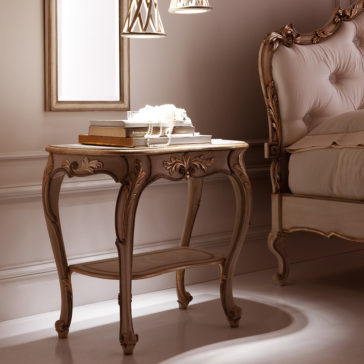 Opulent Carved Italian Bedside Table An elegant vintage Opulent Carved Italian Bedside Table with ornate carvings holds a stack of books and a pearl necklace under soft, warm lighting. It is set next to a similarly styled bed with a tufted headboard and a picture frame in the background.