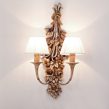 Designer Rococo Bouquet Wall Light The Designer Rococo Bouquet Wall Light exudes vintage charm with its intricate floral and leaf patterns finished in gold. This ornate sconce features two lights, each adorned with a white pleated lampshade. Mounted on a white wall, it casts a warm glow that creates soft shadows around the fixture.