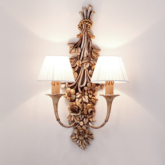 The Designer Rococo Bouquet Wall Light exudes vintage charm with its intricate floral and leaf patterns finished in gold. This ornate sconce features two lights, each adorned with a white pleated lampshade. Mounted on a white wall, it casts a warm glow that creates soft shadows around the fixture.