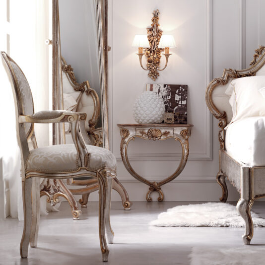 A luxurious bedroom features ornate furniture with gold accents. A carved chair pairs with an elegant mirrored vanity. The Designer Rococo Bouquet Wall Light illuminates a small table beside the bed, holding a decorative lamp and book. A plush rug lies on the floor, enhancing the room's opulent ambiance.