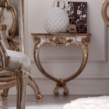 An Ornate Rococo Wall Mounted Bedside Table with a half-circle shape and gold detailing supports a white, textured vase resembling a pineapple. A framed photo and decorative item are also placed on top. The table stands against a white wall near an upholstered chair and a large mirror.