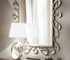 Italian Designer Gold Plated Table Lamp A decorative mirror with an ornate, wavy-edged frame is mounted on the wall. In front of it is the Italian Designer Gold Plated Table Lamp, featuring a white lampshade and a sculptural silver base. The setup is part of a well-lit, modern interior space.
