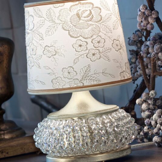 The Small Glamorous Crystal Table Lamp highlights a white floral patterned shade and a base adorned with intricate crystal designs, set against an alloy decorative piece and a branch with dried berries.