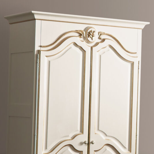 The White Louis Reproduction Two Door Armoire is a cream-colored wooden piece featuring double doors with matching gold handles, ornate gold trim details, and a decorative emblem at the top center. It stands beautifully against a neutral, light brown background.