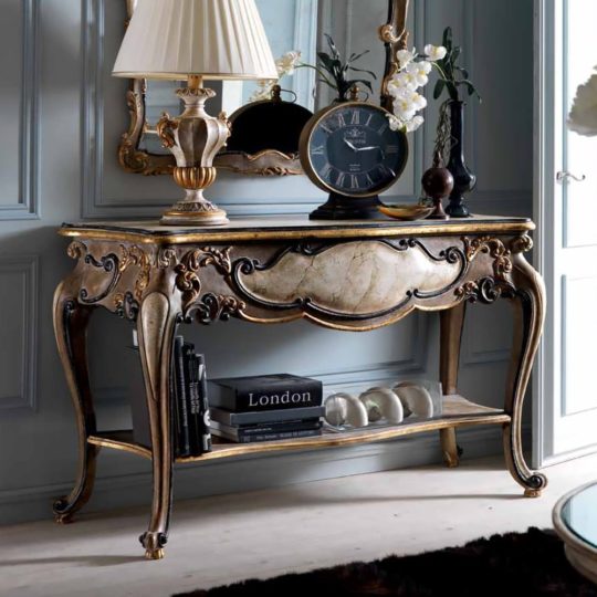 A Classic Italian Antiqued Console Table with ornate gold accents and curved legs displays a lamp, black clock, white flowers in a vase, and books beneath a mirror on an elegant wall.