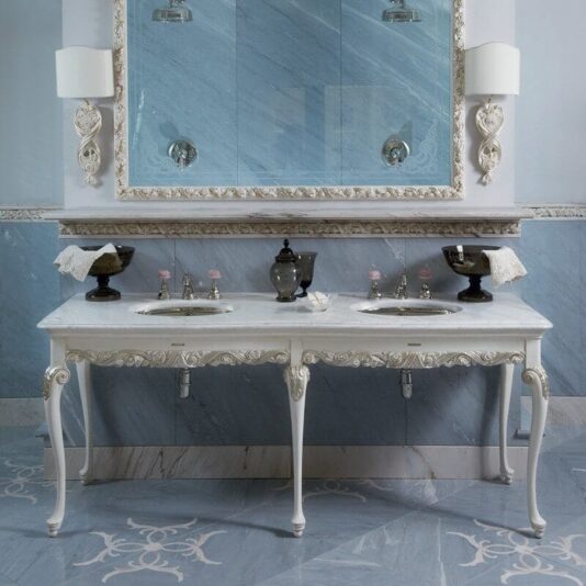 A luxurious bathroom features the Classic Italian Marble Topped Double Bathroom Basin, boasting an ornate white design and two sinks. The large mirror above is framed with intricate detailing, while elegant wall sconces flank either side. Blue marble tiles and a decorative floor pattern complete the opulent look.