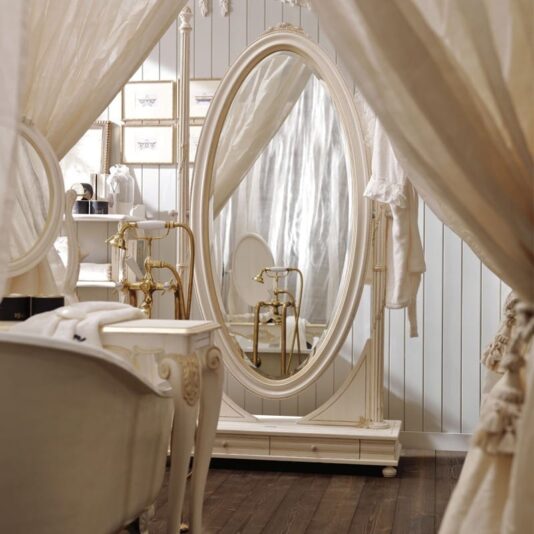 An elegant, vintage-style bathroom showcases a spacious Classic Italian Oval Dressing Mirror adorned with an ornate white frame, complemented by flowing sheer curtains on either side. The dark wooden floor provides a striking contrast to the white walls and furnishings, which include a timeless white bathtub featuring gold fixtures.