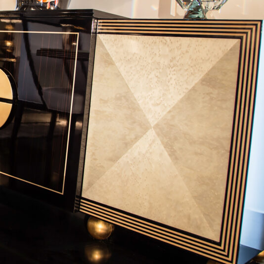 The Contemporary Ebony and Sycamore Veneered Buffet features a sleek, glossy black finish adorned with rectangular and circular gold accents on the left door and a textured beige geometric design bordered by a black and gold frame on the right. This cabinet stands elegantly on gold spherical feet.
