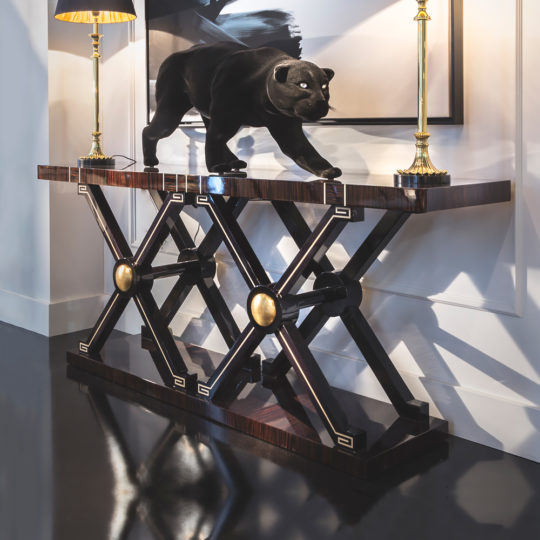 The Contemporary Ebony Criss Cross Design Console Table with gold accents showcases a black panther sculpture and two gold lamps. Set against a white wall, it features X-shaped legs and is topped by large framed artwork above.