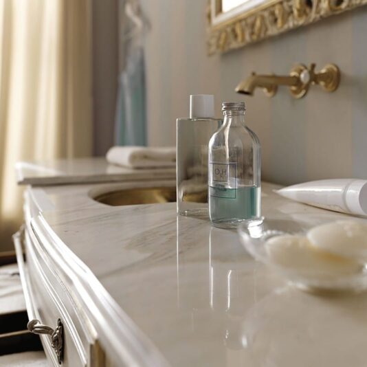 Designer Italian Bathroom Cabinet with Sliding Top A marble bathroom countertop featuring a brass faucet and an ornate mirror. On the countertop are two bottles—one transparent with blue liquid, the other opaque—and a glass dish containing white soap bars. A rolled towel rests near the sink, while in the background stands a Designer Italian Bathroom Cabinet with Sliding Top.